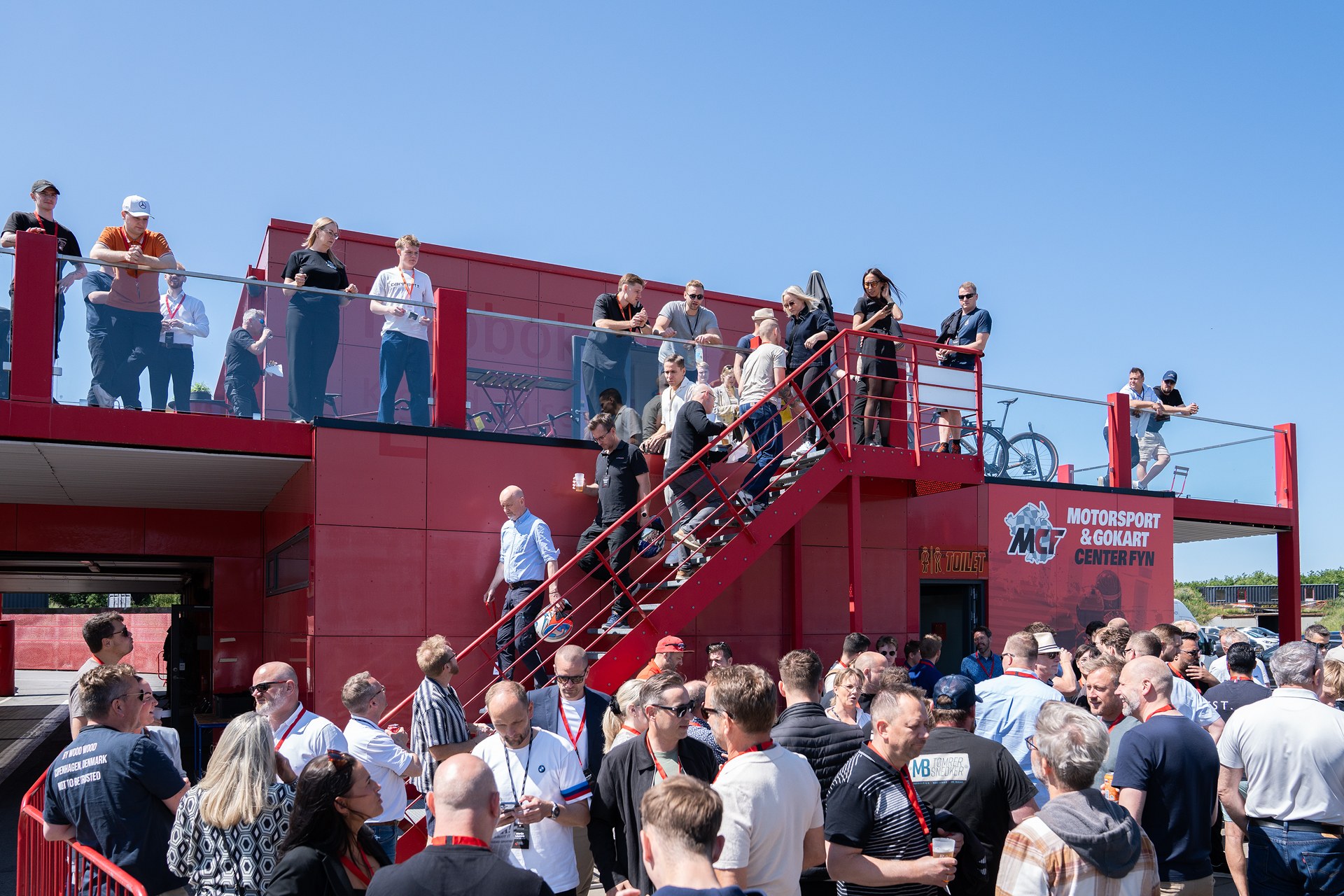 Gæster samlet foran en rød bygning og på altanen over dem til sommerfest